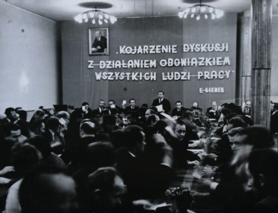 Masa ludzi słucha przemówienia mężczyzny, na ścianie napis 'Kojarzenie dyskusji z działaniem obowiązkiem wszystkich ludzi pracy. E-Giebek'