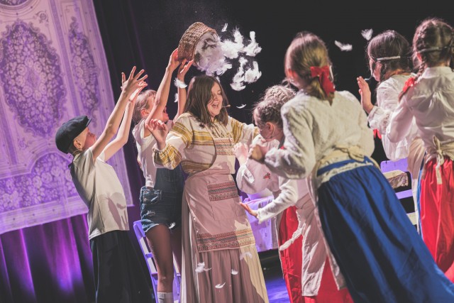 Widok z lewej strony. Dzieci stoją w półkolu wokół dziewczynki w długiej sukience i fartuchu, na którą ktoś właśnie wysypuje kosz pełen piórek. Piórka unoszą się w powietrzu, a wszystkie dzieci śmieją się i wyciągają ręce w górę. 