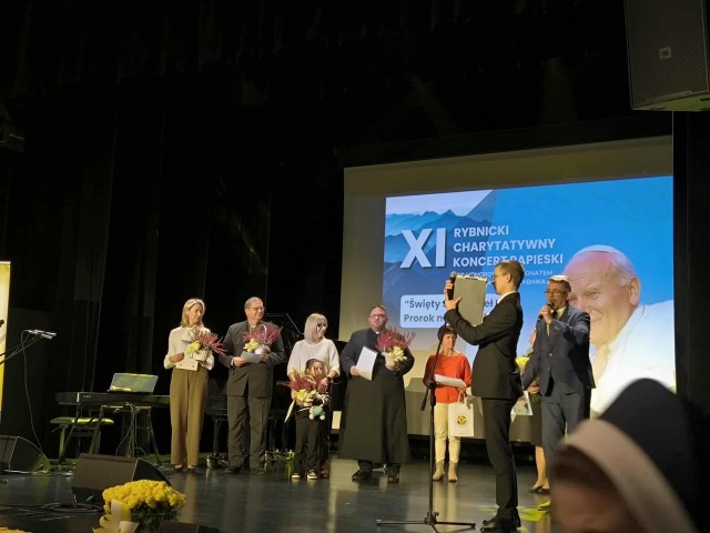 Na scenie podczas XI Rybnickiego Charytatywnego Koncertu Papieskiego stoi grupa osób trzymających kwiaty i dyplomy. W tle widoczny jest ekran z wizerunkiem Jana Pawła II oraz napisem informującym o wydarzeniu. Dwóch mężczyzn przy mikrofonach prowadzi