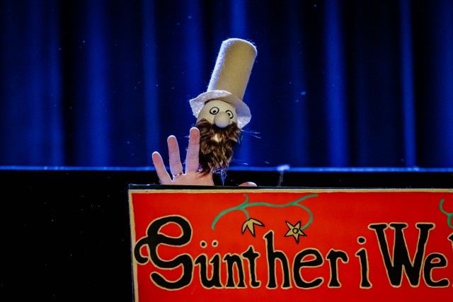 nad czerwonym szyldem widać fragment dłoni z pacynką w kapeluszu. Sytuacja sceniczna