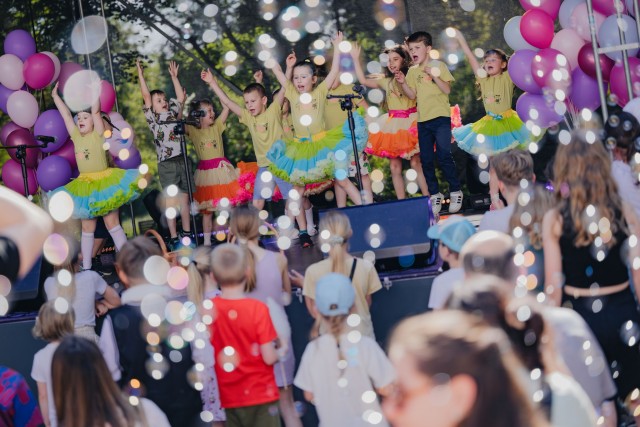 Dzieci tańczą i śpiewają na scenie w parku, ozdobionej balonami, dla publiczności złożonej z ich rówieśników i dorosłych.