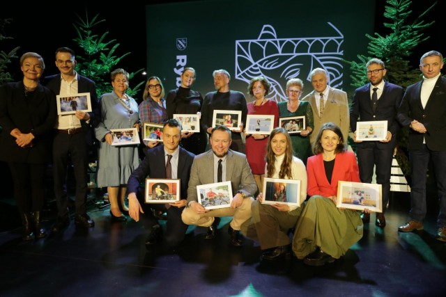 Zdjęcie grupowe na scenie, osoby w różnym wieku pozują z ramkami ze zdjęciami. W tle choinki.