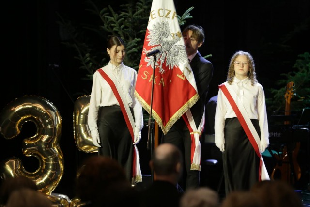 trzy młode osoby w poczcie sztandarowej, mają biało-czerwone wstęgi, osoba w środku trzyma sztandar.