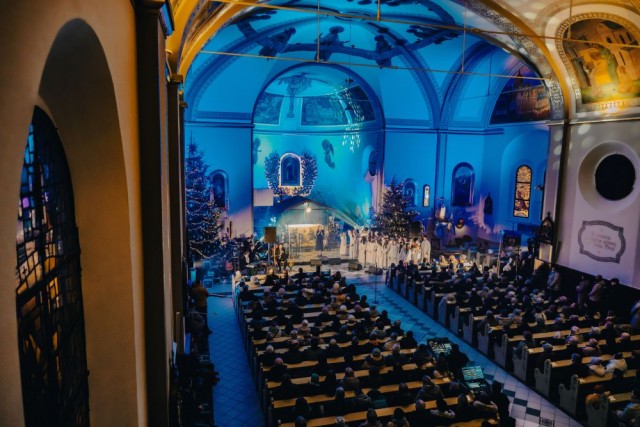 Widok z góry z boku na nawę główną kościoła. Ściany kościoła oświetlone niebieskim, nastrojowym światłem. W centrum (ołtarz): zespół, grupa osób w białych strojach. W kościelnych ławkach siedzą ludzie.