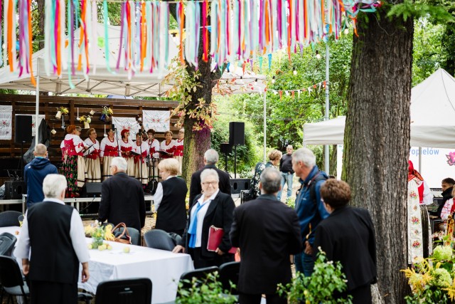 Wydarzenie plenerowe w ogrodzie, który został przystrojony kolorowymi wstążkami. Na scenie występuje kobiecy zespół ludowy, przy stole stoi grupa starszych osób i patrzy na scenę. W tle namiot.
