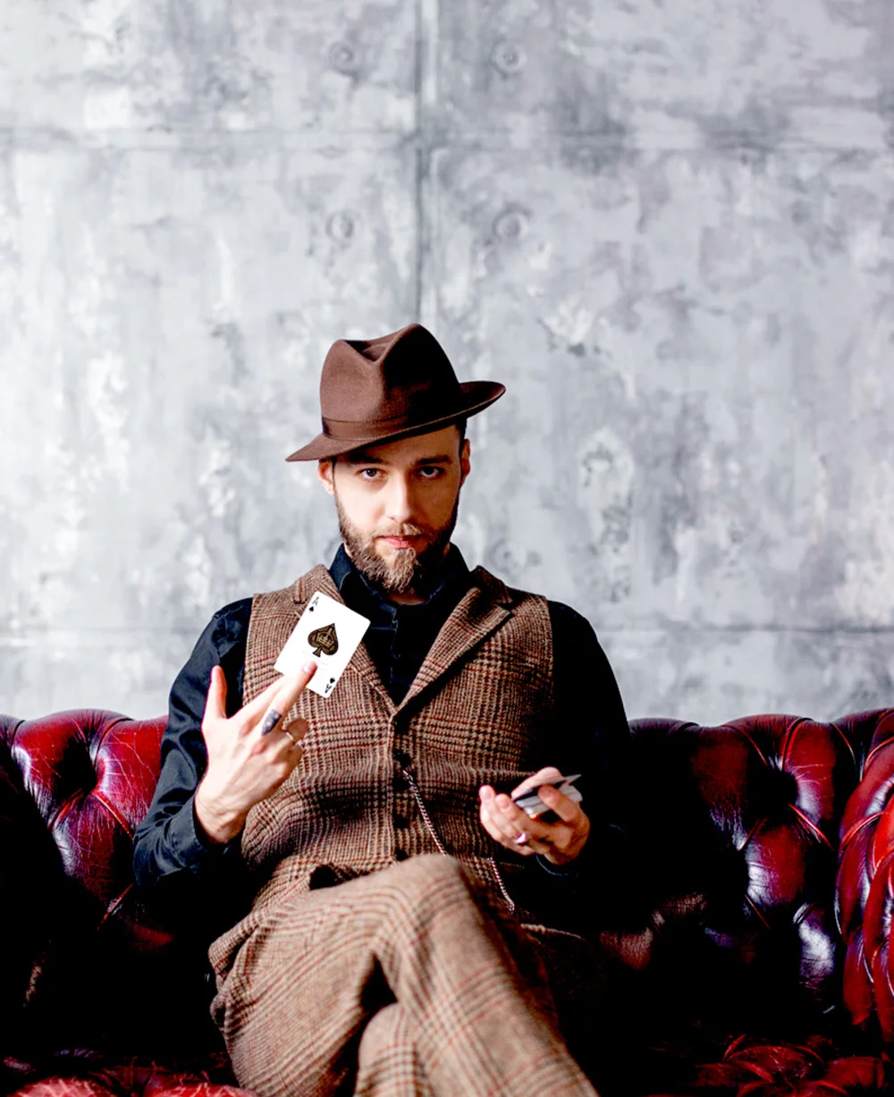 mężczyzna w kapeluszu siedzi na kanapie z talią kart z w jednej ręce i asem w drugiej