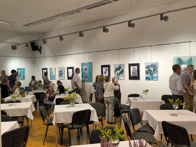 Widok na salę, część osób siedzi przy stolikach, część ogląda wystawę zawieszoną na ścianach.